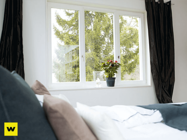 Helles Schlafzimmer mit großem Fenster, Bett, Vorhängen und Zimmerpflanze auf der Fensterbank.
